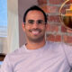 A smiling man wearing glasses and a dark t-shirt with a logo, standing against a neutral background.