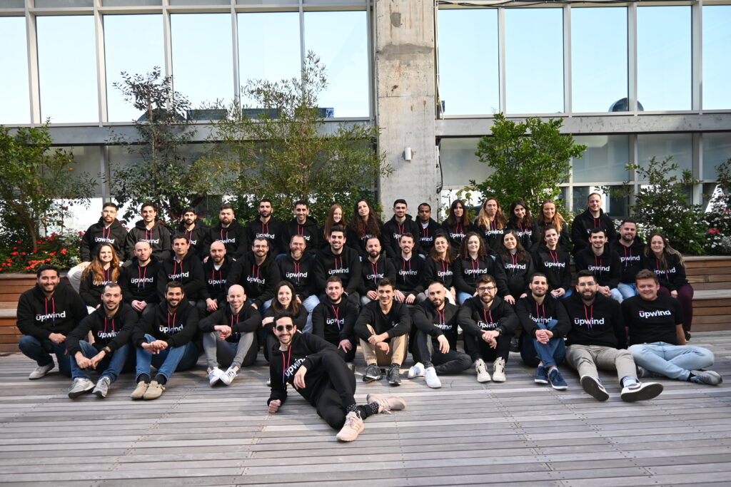 A group of people wearing matching black Upwind shirts are seated and standing on wooden decking with plants and a large windowed building in the background. They are posing for a group photo outdoors.