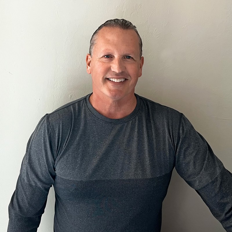 A smiling man with short hair stands in front of a plain light-colored wall, wearing a long-sleeve dark gray shirt.