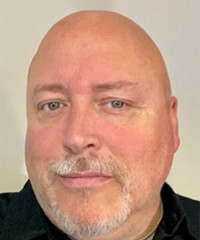 A bald man with a light beard and mustache, wearing a black shirt, looks directly at the camera with a neutral expression. The background is plain and light-colored.