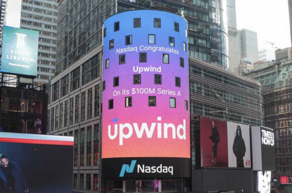 A Nasdaq billboard in Times Square, New York, displays a congratulatory message for Upwind on securing its $100M Series A funding. The background is a gradient of pink to blue. Buildings and advertisements are visible in the surrounding area.