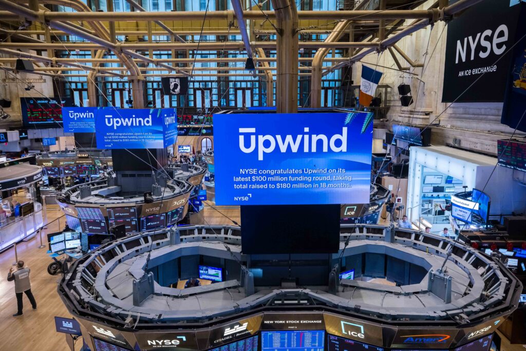 View of the New York Stock Exchange trading floor with multiple screens displaying Upwind. The main screen highlights a congratulatory message for Upwinds latest funding round. Traders desks and electronic equipment are visible.