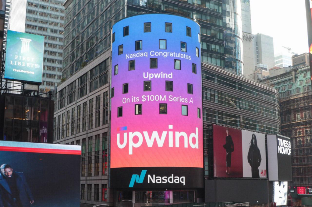 A digital billboard displays, Nasdaq congratulates Upwind on its $100M Series A, with a colorful background. The Nasdaq logo is shown at the bottom. The sign is in a busy urban area with tall buildings.