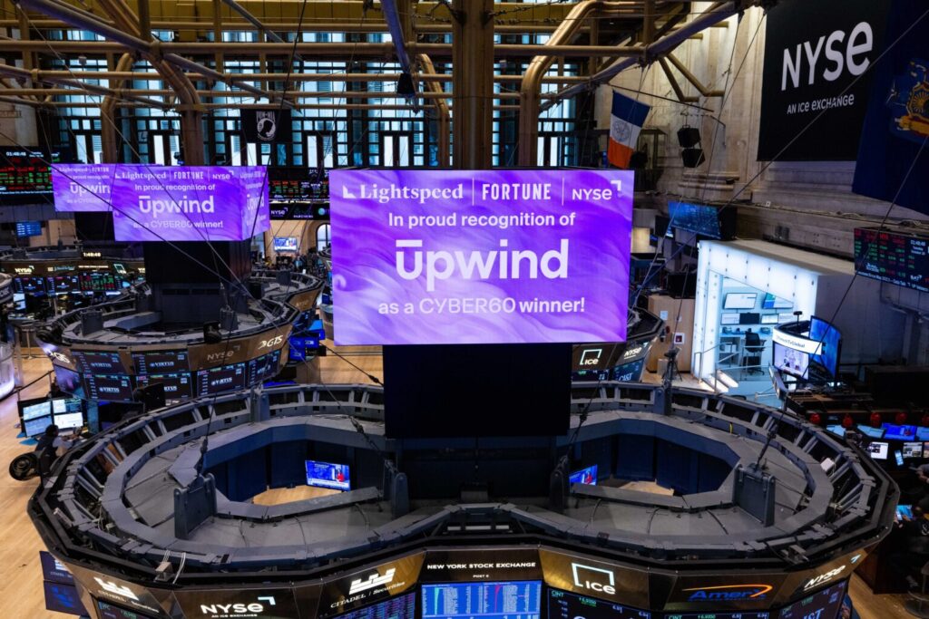 A large screen at the NYSE displays a message: Lightspeed, Fortune, NYSE. In proud recognition of Upwind as a CYBER60 winner! The trading floor is bustling with electronic displays and stock exchange activity.