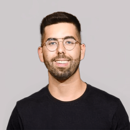 A young man with short dark hair and a beard smiles at the camera. He is wearing round glasses and a black shirt. The background is plain and light gray.