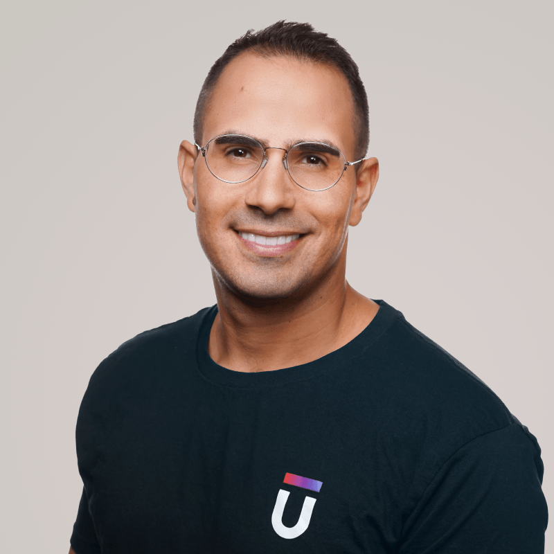 A smiling man with short hair and glasses wears a dark T-shirt featuring a stylized U logo. The background is a plain, light color.