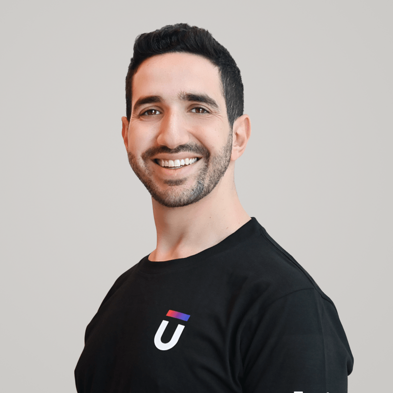 Smiling person with short dark hair and a beard, wearing a black T-shirt with a small logo featuring a red and blue line above the letter U, against a plain light gray background.