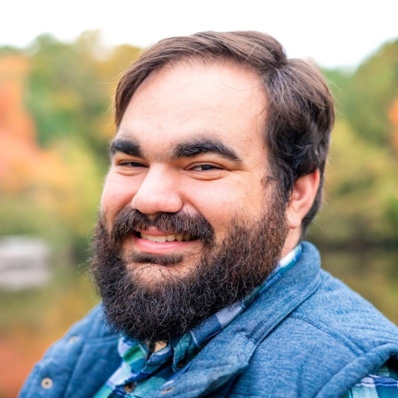 Bearded man smiling outdoors, wearing a blue vest over a plaid shirt. The background features blurred autumn foliage.