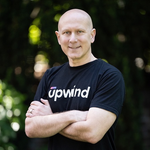 A person stands outdoors with arms crossed, wearing a black t-shirt with the word upwind in white letters. The background is blurred with green foliage. The person is smiling and looking at the camera.